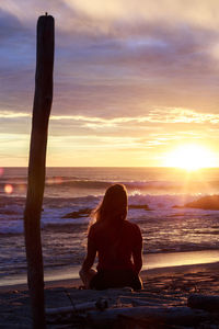 Rear view of woman sitting on sand at beach against sky during sunset