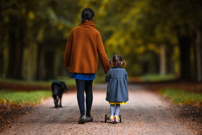 Mother is helping her toddler daughter on the scooter while they are walking their dog in the forest