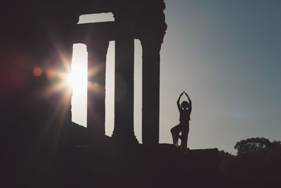 Silhouette mid adult woman exercising at old ruin against sky during sunset