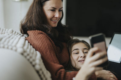 Girl sharing smart phone with smiling mother while lying on sofa in living room