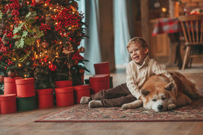 Candid authentic happy little boy in knitted beige sweater hugs dog with bow tie at home on xmas