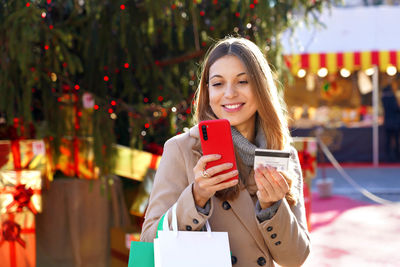 Beautiful smiling girl paying her gifts with mobile phone with christmas markets on the background.