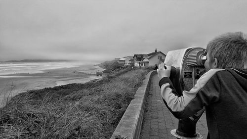 Rear view of boy looking through coin-operated binoculars on promenade against cloudy sky