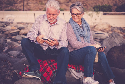 Couple using mobile phones while sitting on rocks at beach