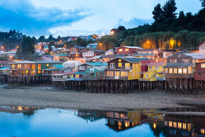 Illuminated buildings by lake against sky at dusk