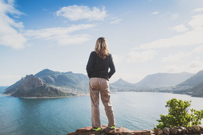 Rear view of woman standing on stone retaining wall while looking at hout bay against sky