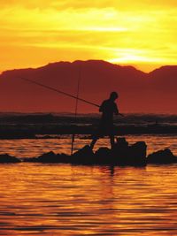 Silhouette man fishing in sea against orange sky