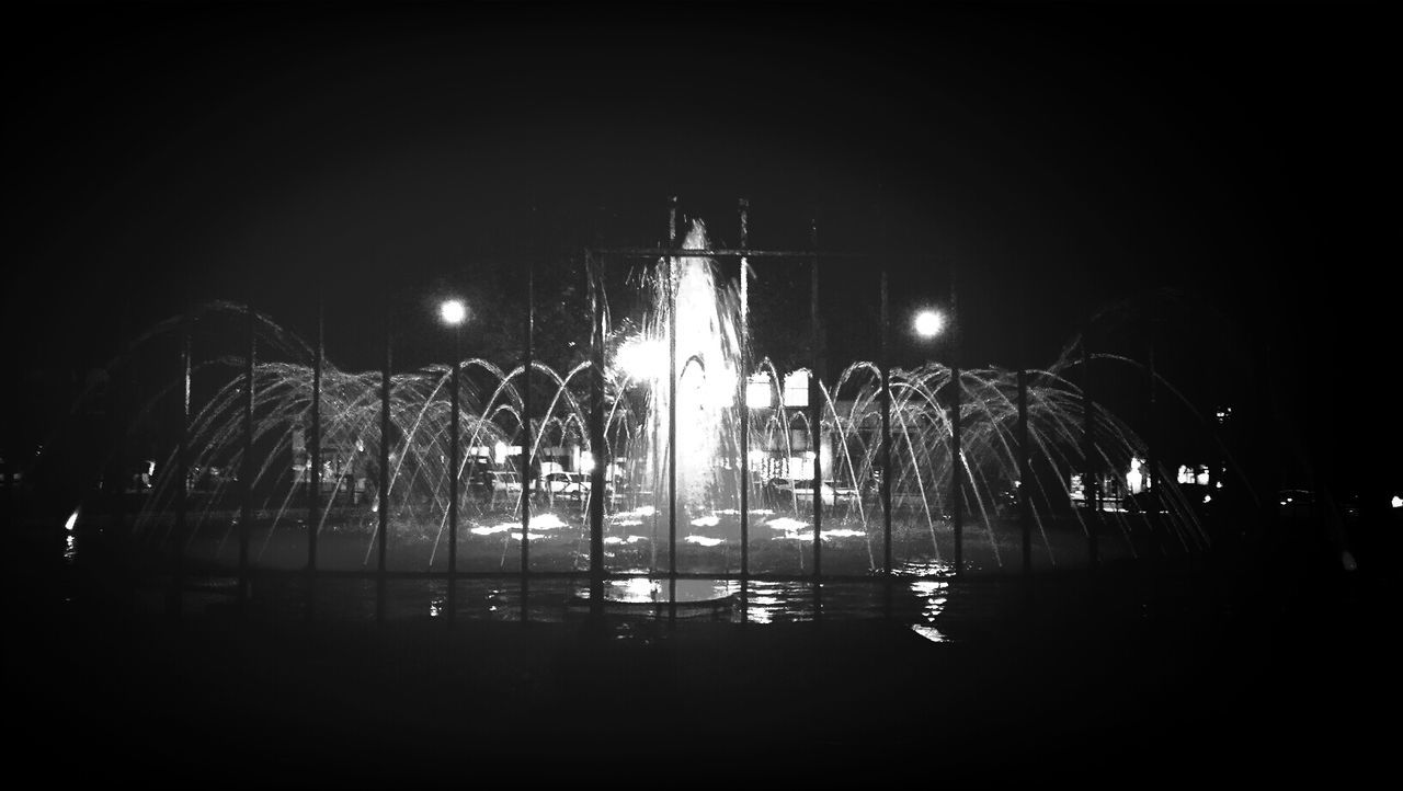 night, illuminated, water, reflection, lighting equipment, street light, dark, long exposure, sky, glowing, clear sky, copy space, motion, outdoors, light - natural phenomenon, fountain, building exterior, city, tree, built structure