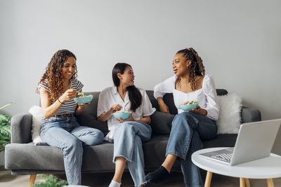 Smiling young multiracial roommates eating salad together sitting on sofa at home