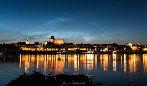 Illuminated buildings by river against sky at night