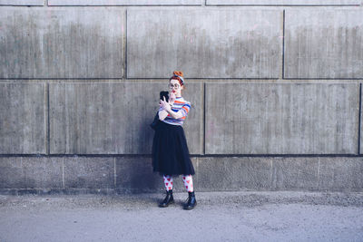 Young woman looking at mobile phone while applying lipstick against wall