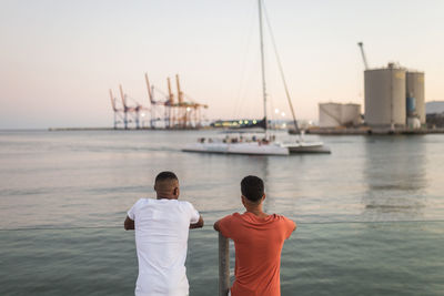 Back view of anonymous young ethnic friends spending time together on river embankment in evening