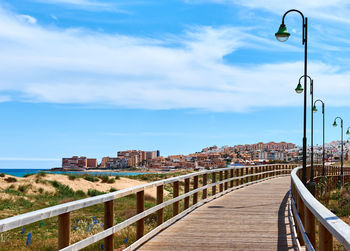 Wooden footbridge by city against cloudy sky