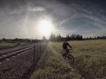 Man cycling on field by railroad tracks against sky during sunny day
