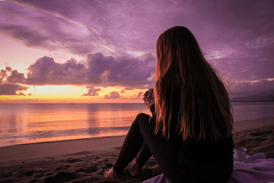 Young woman sitting at beach against cloudy sky during sunset