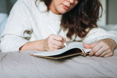 Young adult brunette woman forty in white knitted sweater writes notes in planner on bed at home