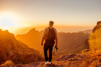 Rear view of man holding camera while standing on mountain against sky during sunset