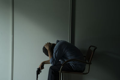 Side view of depressed man holding gun while sitting on chair against wall