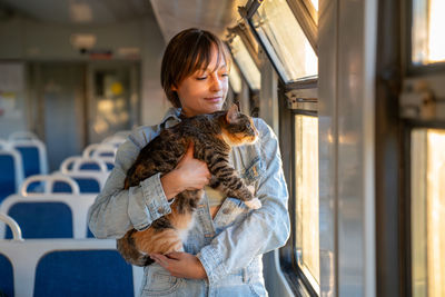 Smiling woman with fluffy tricolor cat traveling riding in electric train together at setting sun.