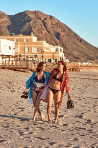 Two girls smiling as they walk on the sand of the beach in their swimming costumes with their shoes