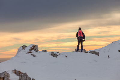 Rear view of man standing on snowcapped mountain against sky during sunset
