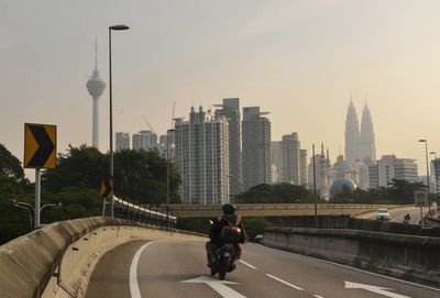 Person riding motorcycle on road by menara kuala lumpur tower and petronas towers against sky