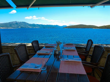 Empty chairs and tables by sea against sky