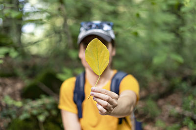 Young woman in defocus holding autumn fallen yellow leaf in front of face, faceless concept