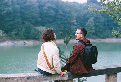 Couple talking while sitting on railing against lake and forest