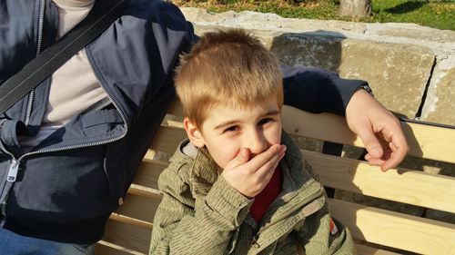 Portrait of cute boy covering mouth while sitting by father on bench