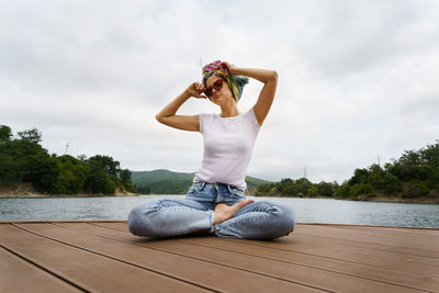 Full length of young woman sitting by lake against sky