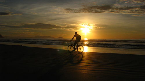 Man riding bicycle at beach against sky during sunset