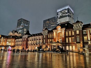Illuminated buildings by wet street against sky in city at night