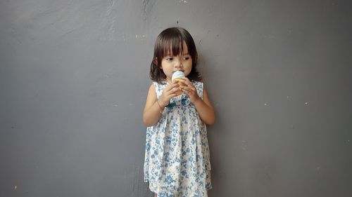 Girl eating ice cream while standing against gray wall