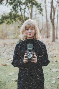 Portrait of young woman holding camera while standing on field