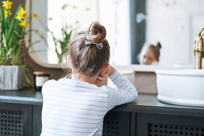 Cute bored little girl at bright bathroom at home, people from behind