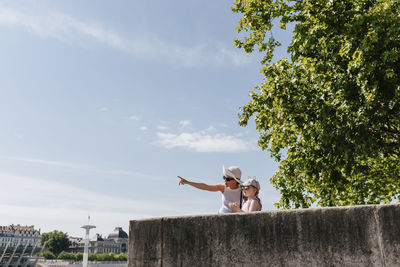 Low angle view of mother gesturing while standing with daughter against sky in city during sunny day