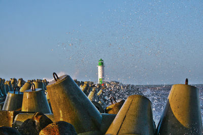 Wave splash on groyne against a lighthouse and clear sky