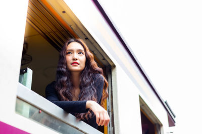 Portrait of young woman looking away while sitting on window