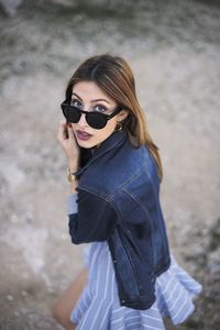 From above of cool young female wearing stylish casual clothes and sunglasses standing in nature and touching hair looking at camera