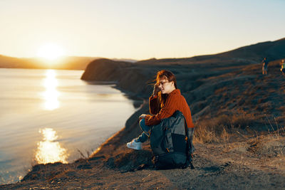 Man sitting on rock at shore against sky during sunset