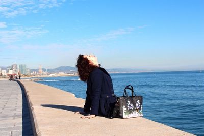 Side view of woman sitting on retaining wall by sea against sky