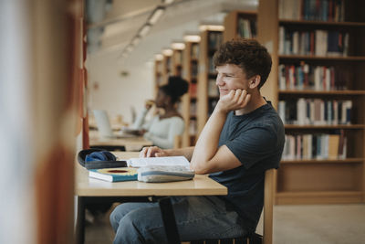 Side view of smiling student sitting with hand on chin in library