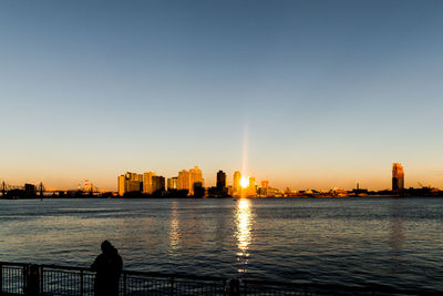 Silhouette city by sea against clear sky during sunset