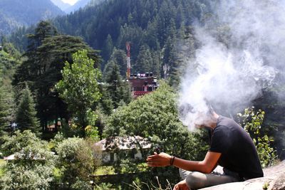 Side view of man smoking cigar while sitting on rock against trees