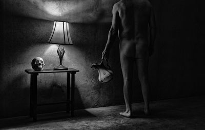 Rear view of naked man standing by illuminated lamp at home