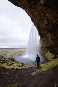 Back view of anonymous male traveler in warm outfit standing in rocky cave and admiring picturesque rapid seljalandsfoss waterfall under cloudy sky in iceland