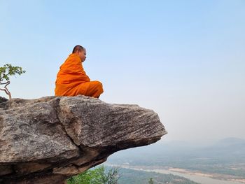 Rear view of man sitting on rock against clear sky