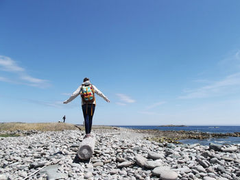 Rear view of woman standing on driftwood at beach against blue sky
