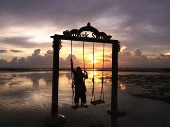 Silhouette woman standing on swing at beach against sky during sunset